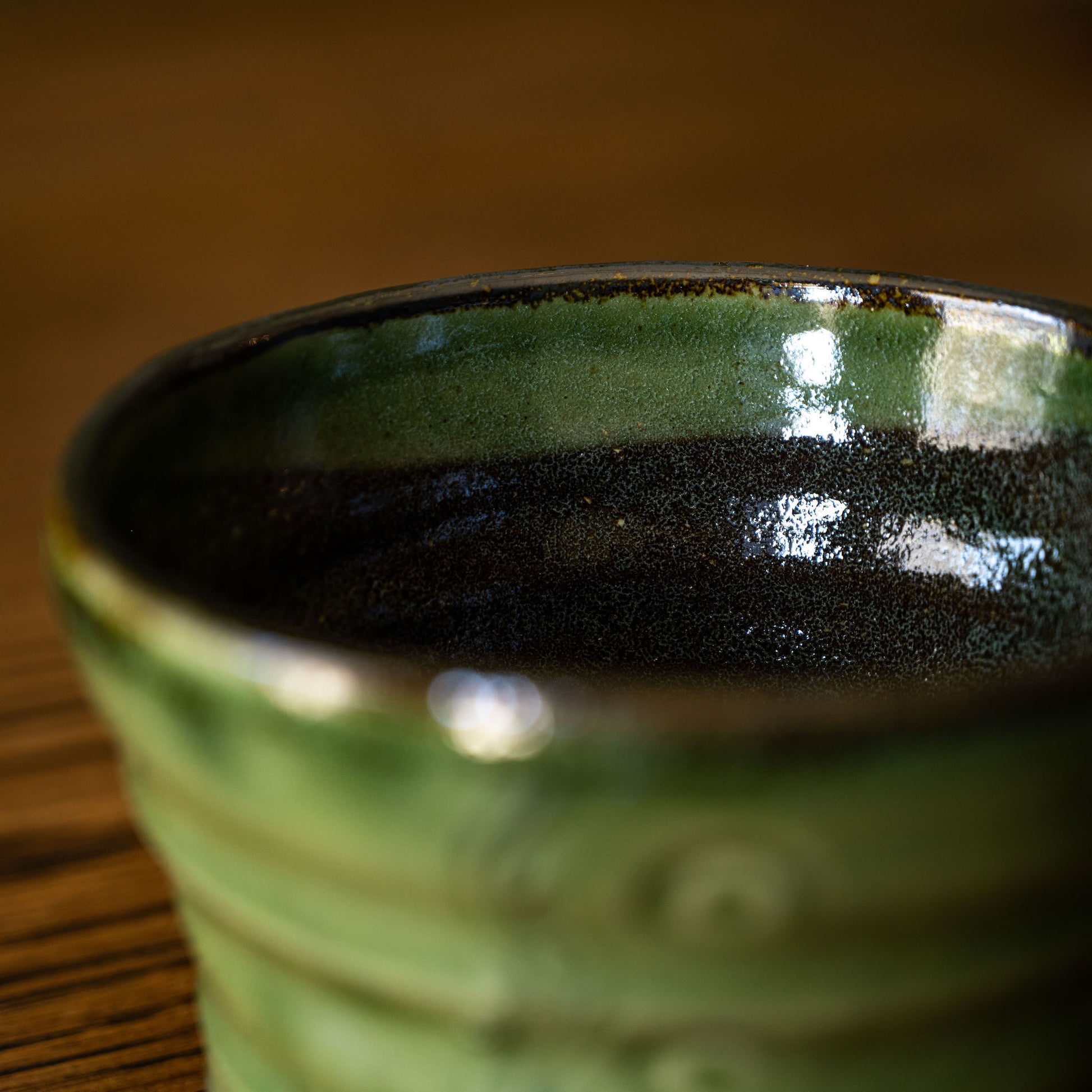 Close-up of Joshua Williams handmade ceramic tea bowl with a textured green glaze and a dark, almost black interior, resting on a wooden surface.
