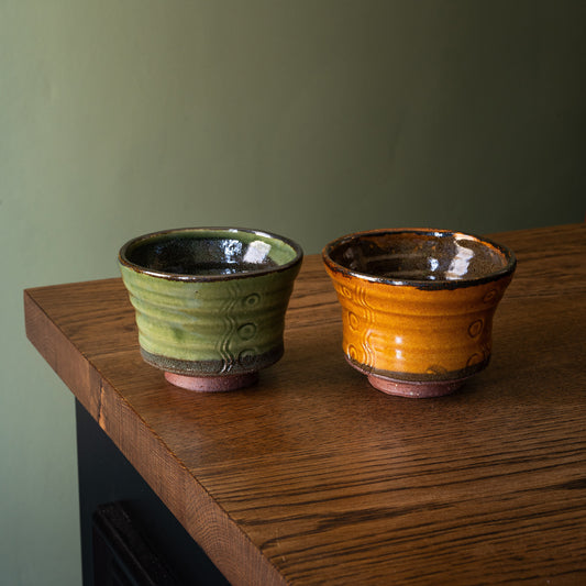 Two handmade Joshua Williams earthenware tea bowls with glossy glazes, one in olive green and one in mustard yellow, sitting on a rustic wooden table against a muted green wall.
