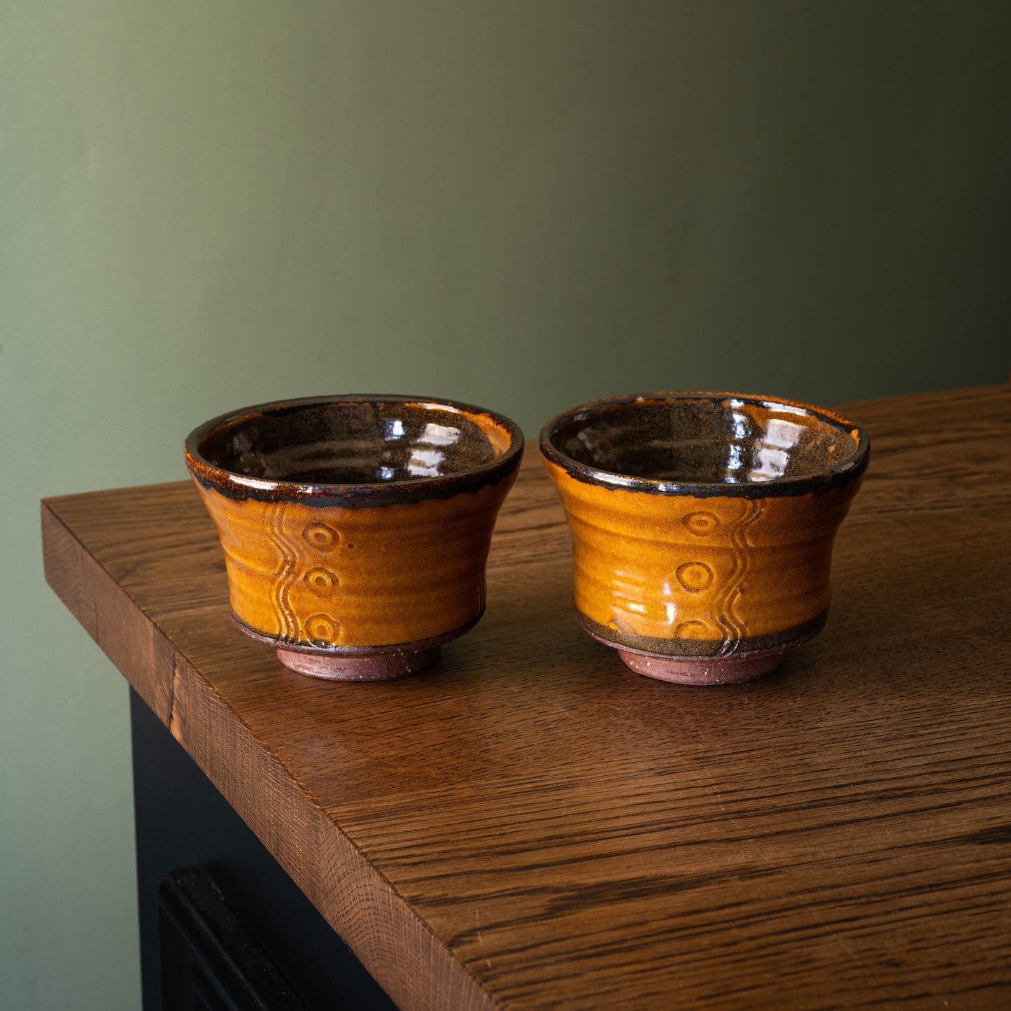 Two handmade Joshua Williams earthenware tea bowls with a glossy amber-orange glaze and dark brown rim, sitting on a rustic wooden table against a muted green wall.