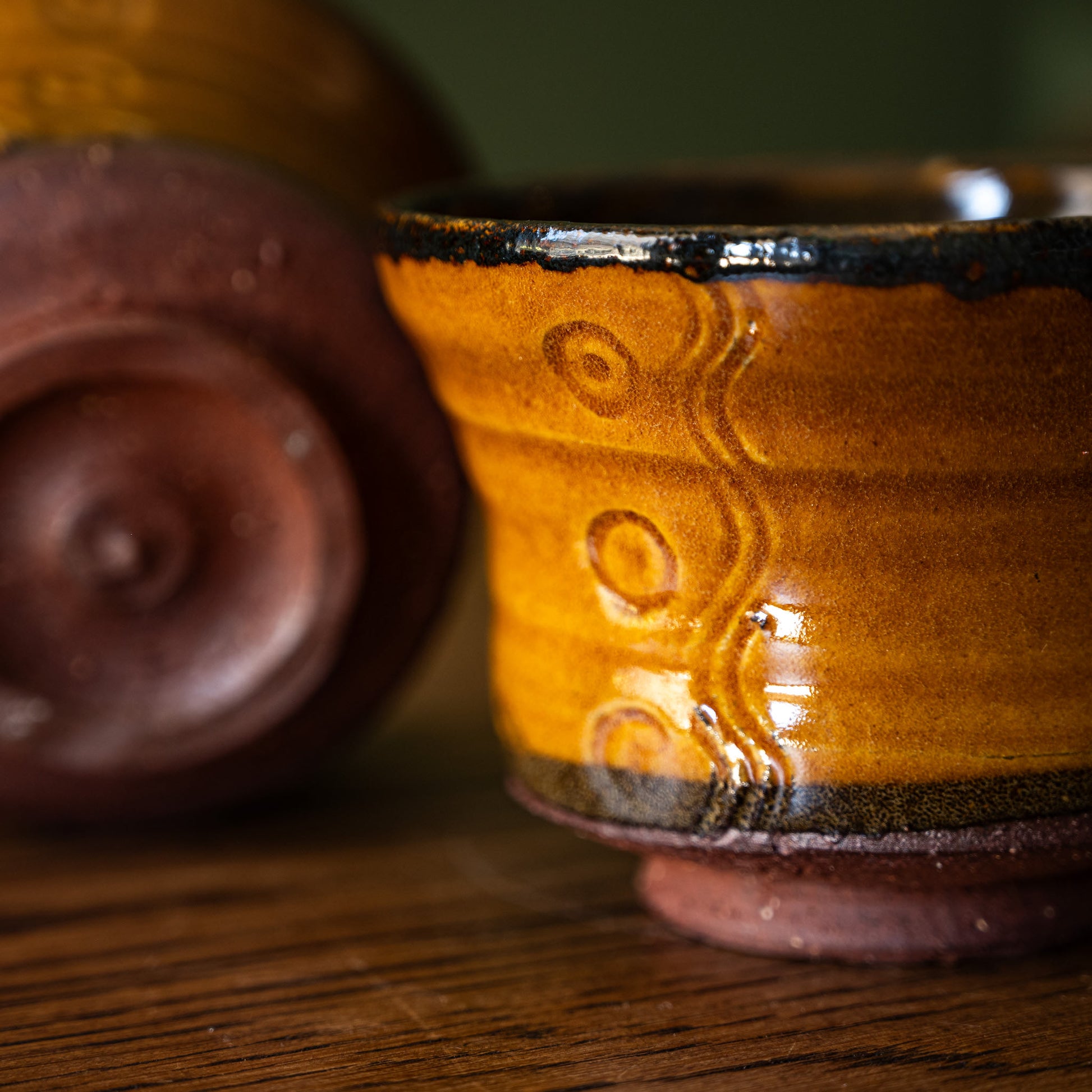 A close-up of Joshua Williams handmade eartenware tea bowl with a glossy amber-brown glaze, featuring embossed circular patterns and a dark rim, set on a wooden surface with another blurred cup in the background.