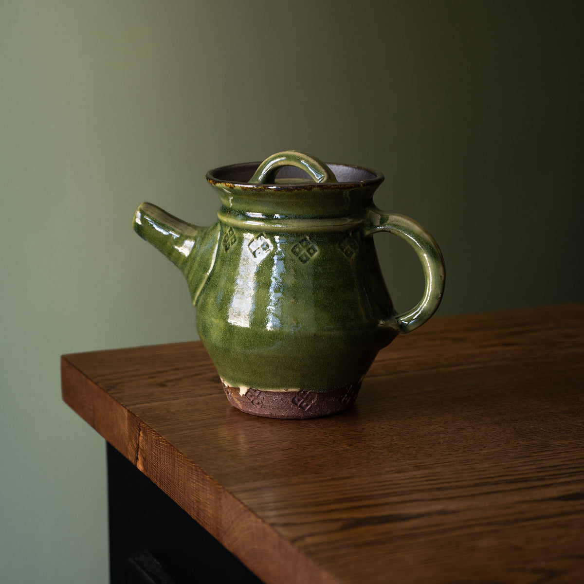 Joshua Williams handmade earthenware teapot with a glossy dark olive green glaze and a rustic brown clay base, resting on a wooden table against a muted green background.
