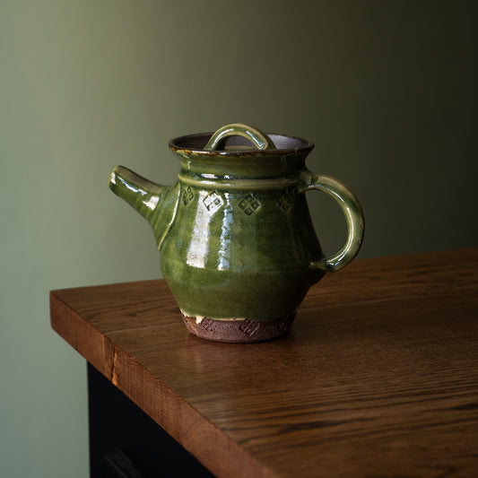 Joshua Williams handmade earthenware teapot with a glossy dark olive green glaze and a rustic brown clay base, resting on a wooden table against a muted green background.