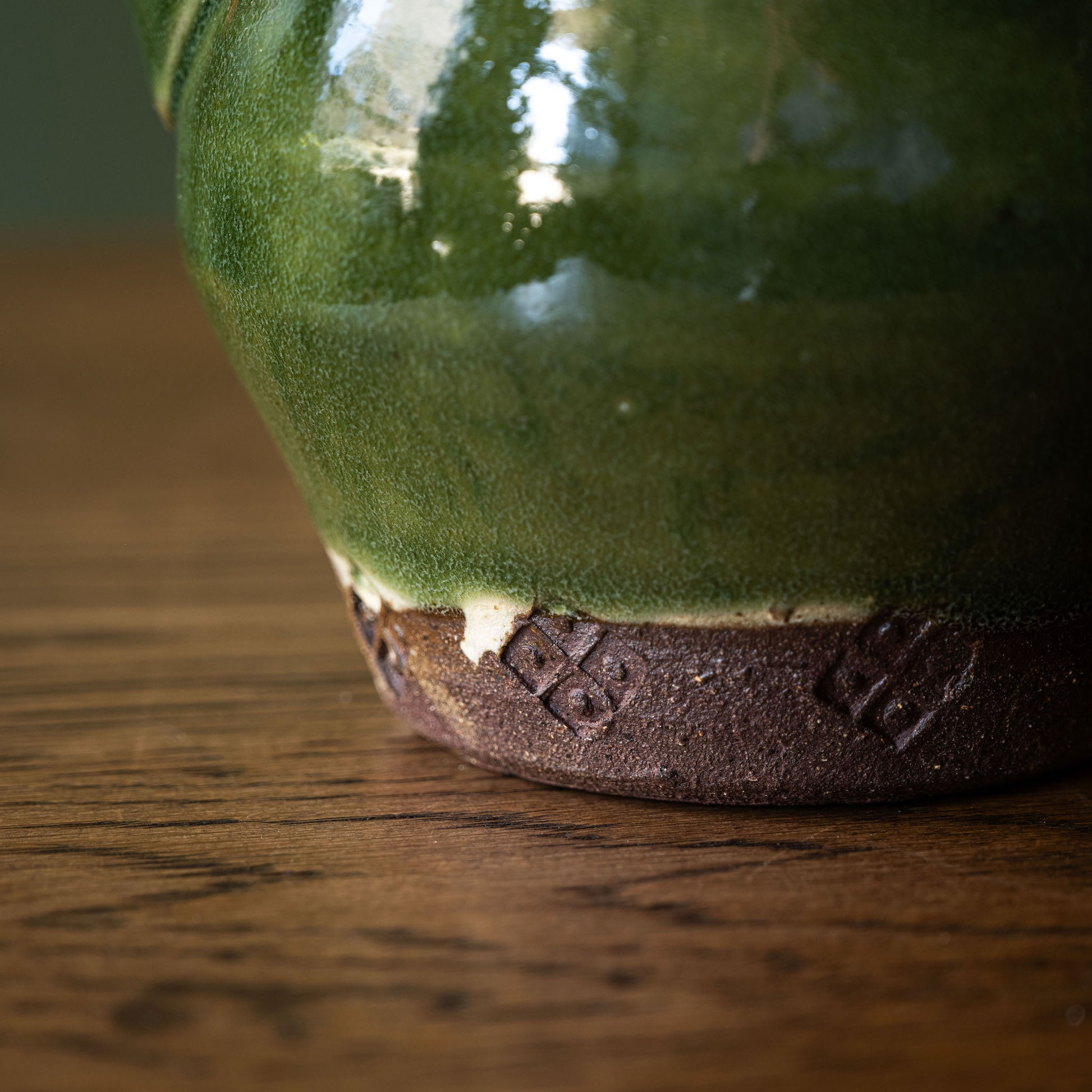 Close-up of Joshua Williams handmade earthenware teapot with a glossy green glaze and a textured brown unglazed base, resting on a wooden surface.