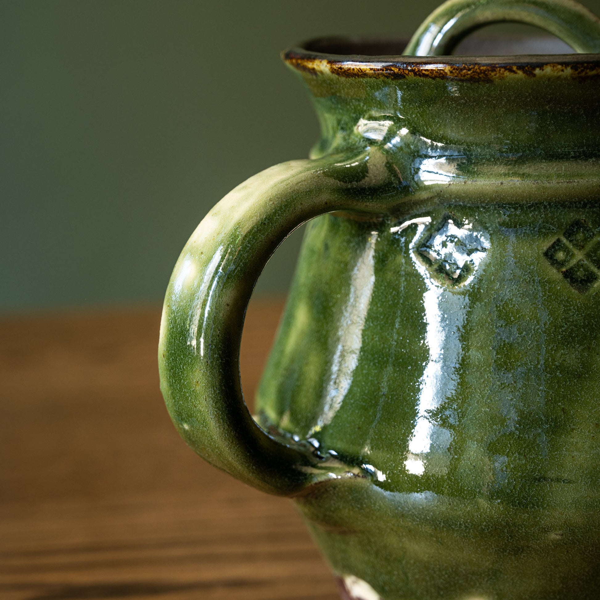 Close-up of Joshua Williams dark green earthenware teapot handle and small embossed diamond motifs around the body, set on a wooden surface against a muted green background.