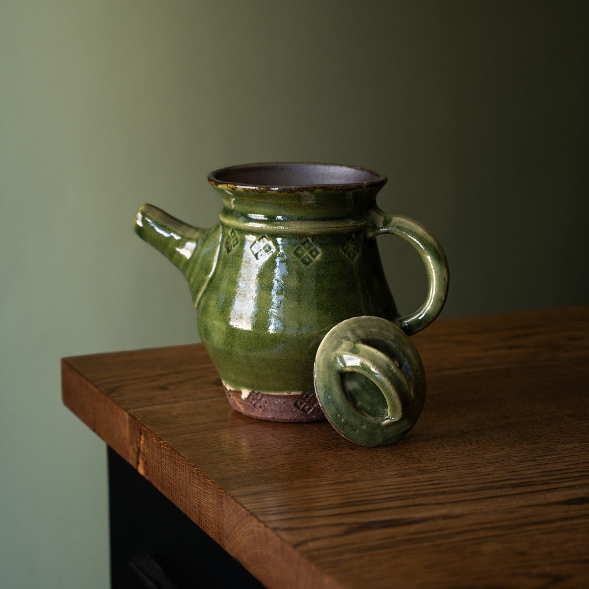 Joshua Williams dark green glazed earthenware teapot with the matching lid resting beside it on a wooden table against a muted green wall.