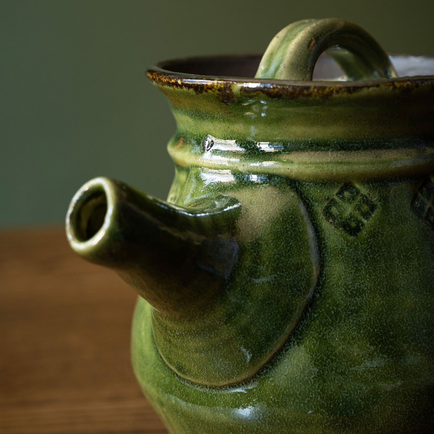 Close-up of Joshua Williams dark green earthenware teapot with a rounded body, short spout and loop handle, photographed on a wooden surface against a muted green background.