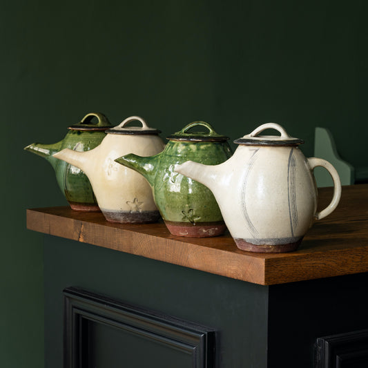 Four Joshua Williams earthenware teapots in green and white on a wooden shelf against a dark green wall.