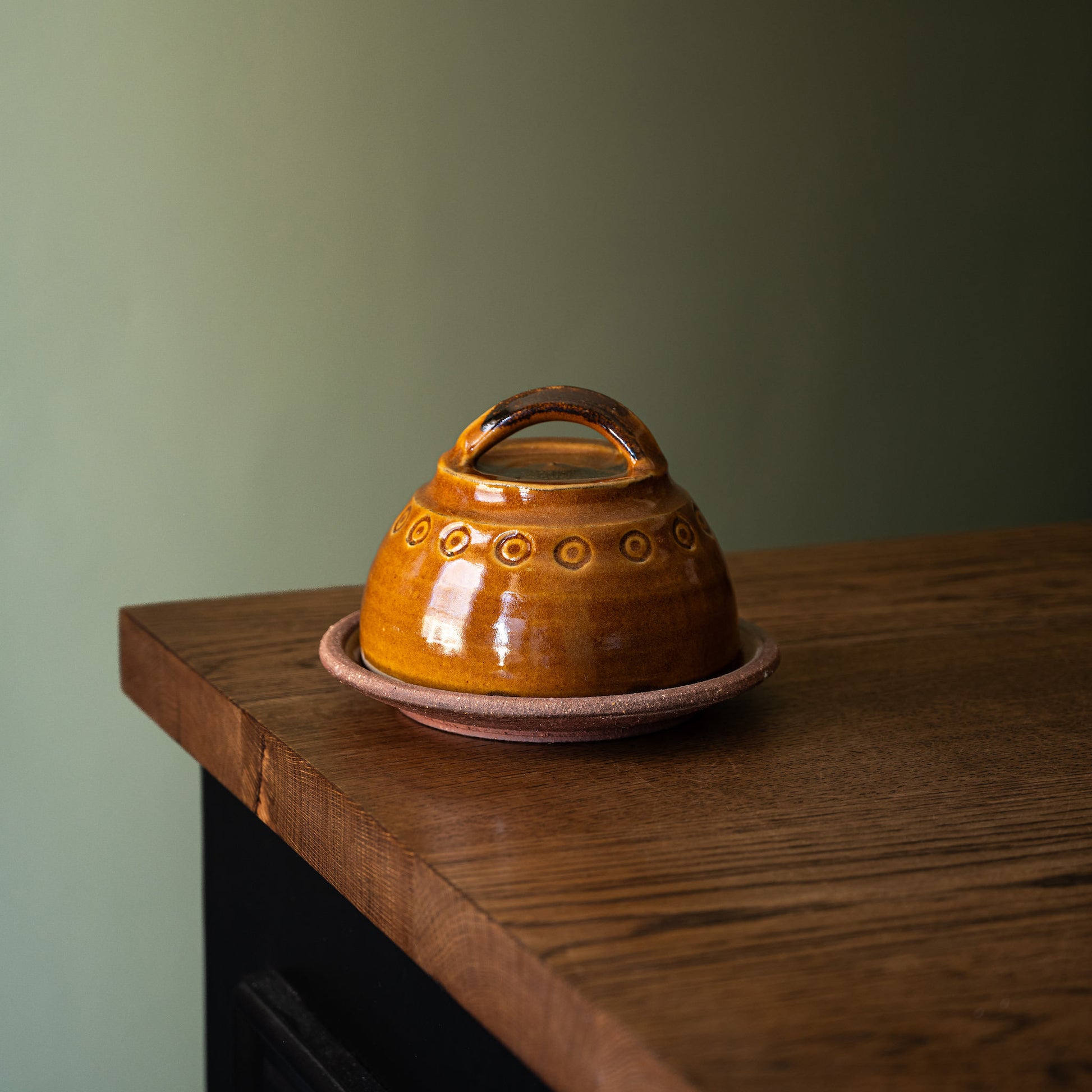 Joshua Williams handmade earthenware two piece domed butter dish with a loop handle with a matching saucer on a wooden tabletop against a muted green wall.