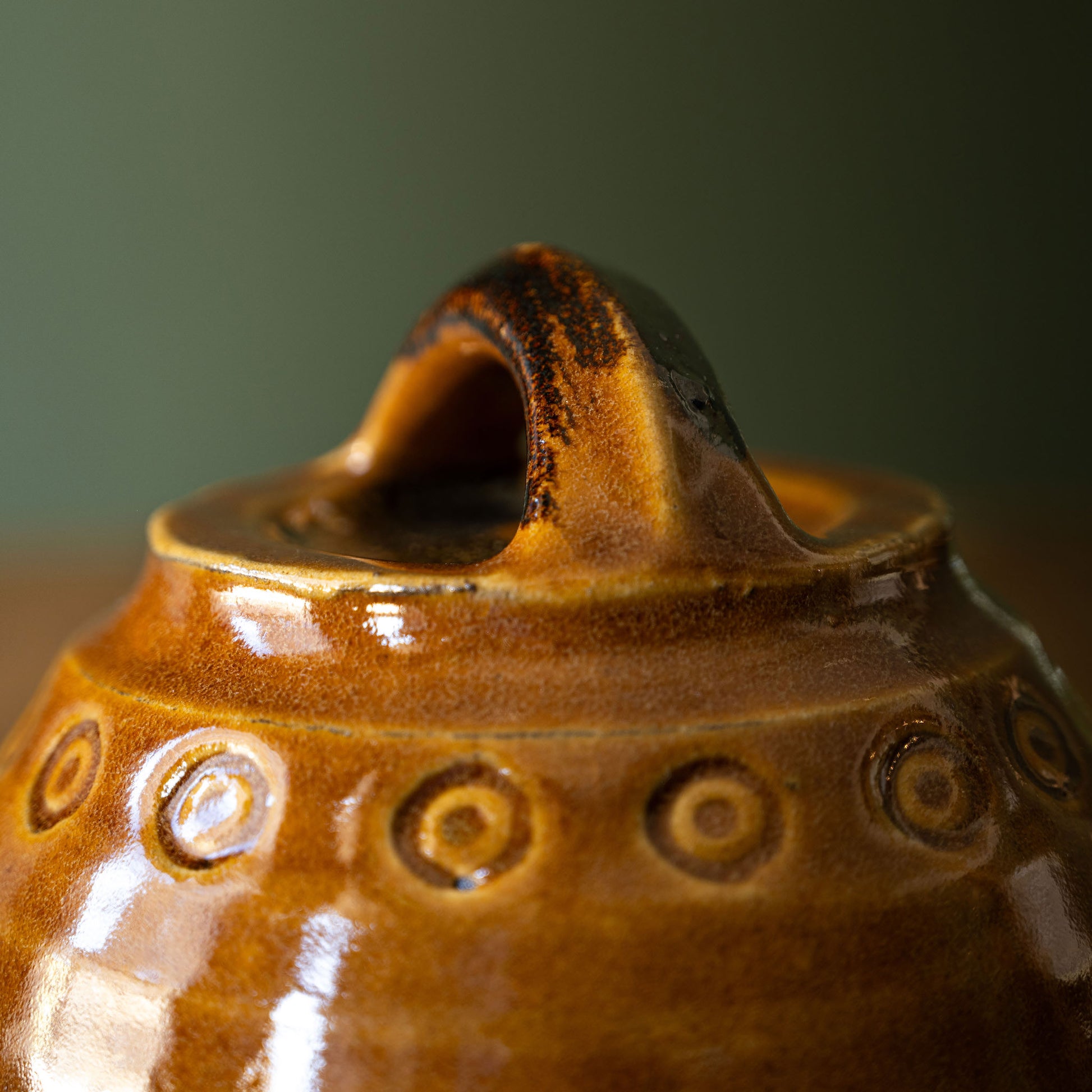 Close-up of a Joshua Williams brown earthenware butter dish with a rounded handle and embossed circular pattern around the neck, set against a soft dark green background.