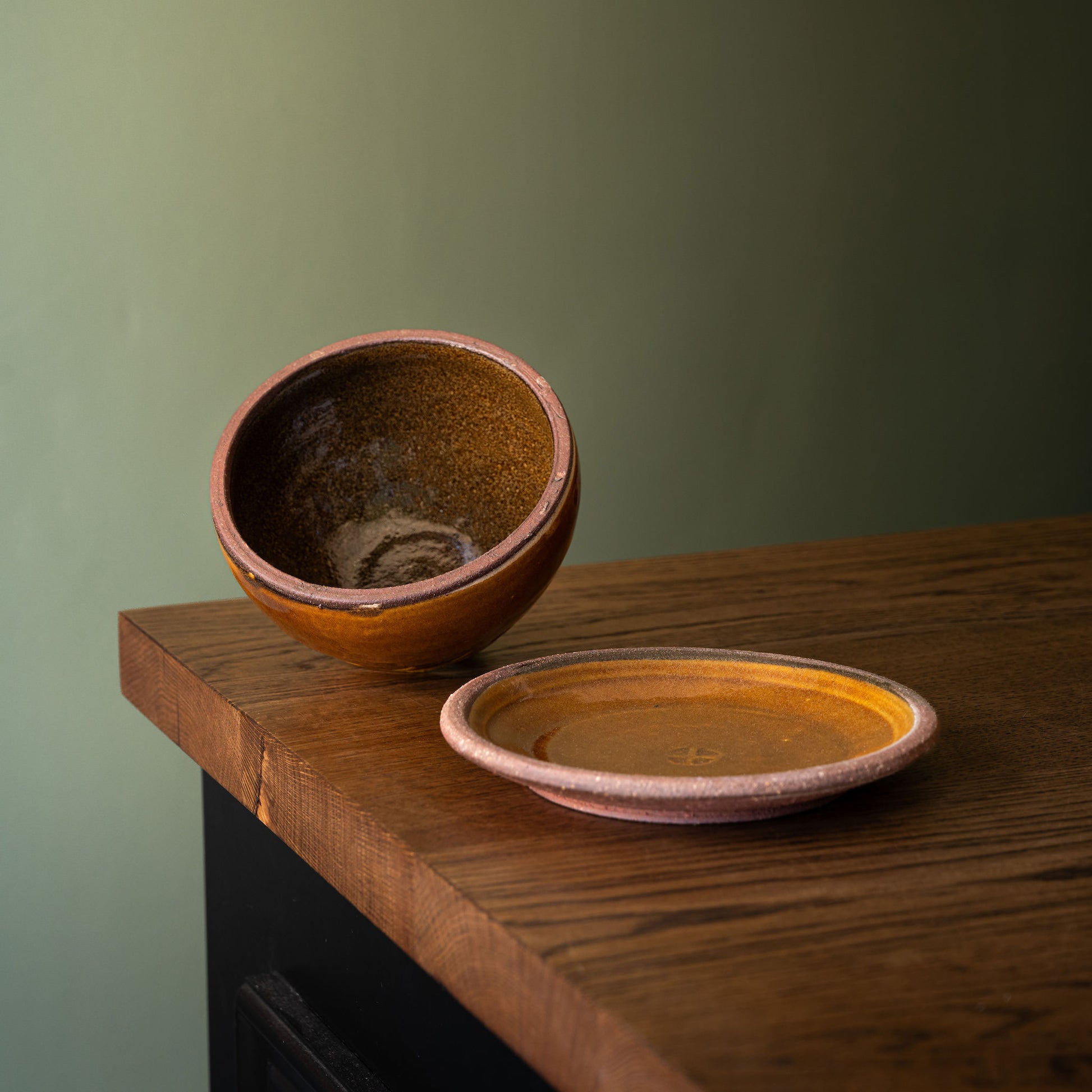 A handmade Joshua Williams  butter dish and matching saucer, displayed on a rustic wooden table against a muted sage green background. The bowl is tipped on its side to show the glazed interior, which features a rich amber-brown colour with subtle speckling, while the saucer rests in front showing a warm ochre glaze and a dusty terracotta rim.