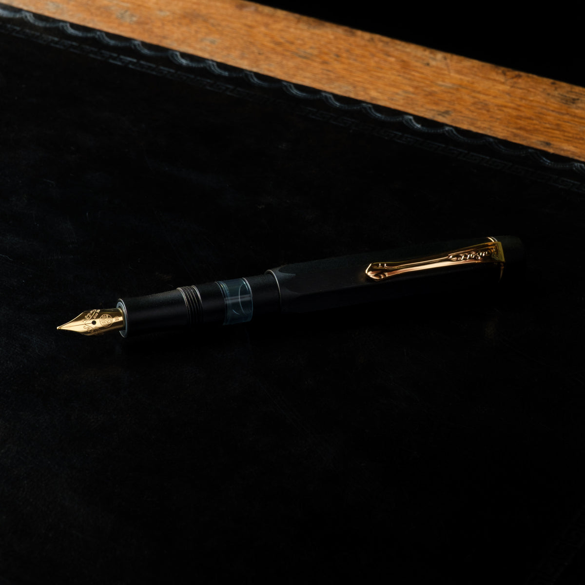 Kaweco Piston Sport AL black fountain pen with gold trim and a gold nib, resting on a black textured surface with a wooden edge in the background.