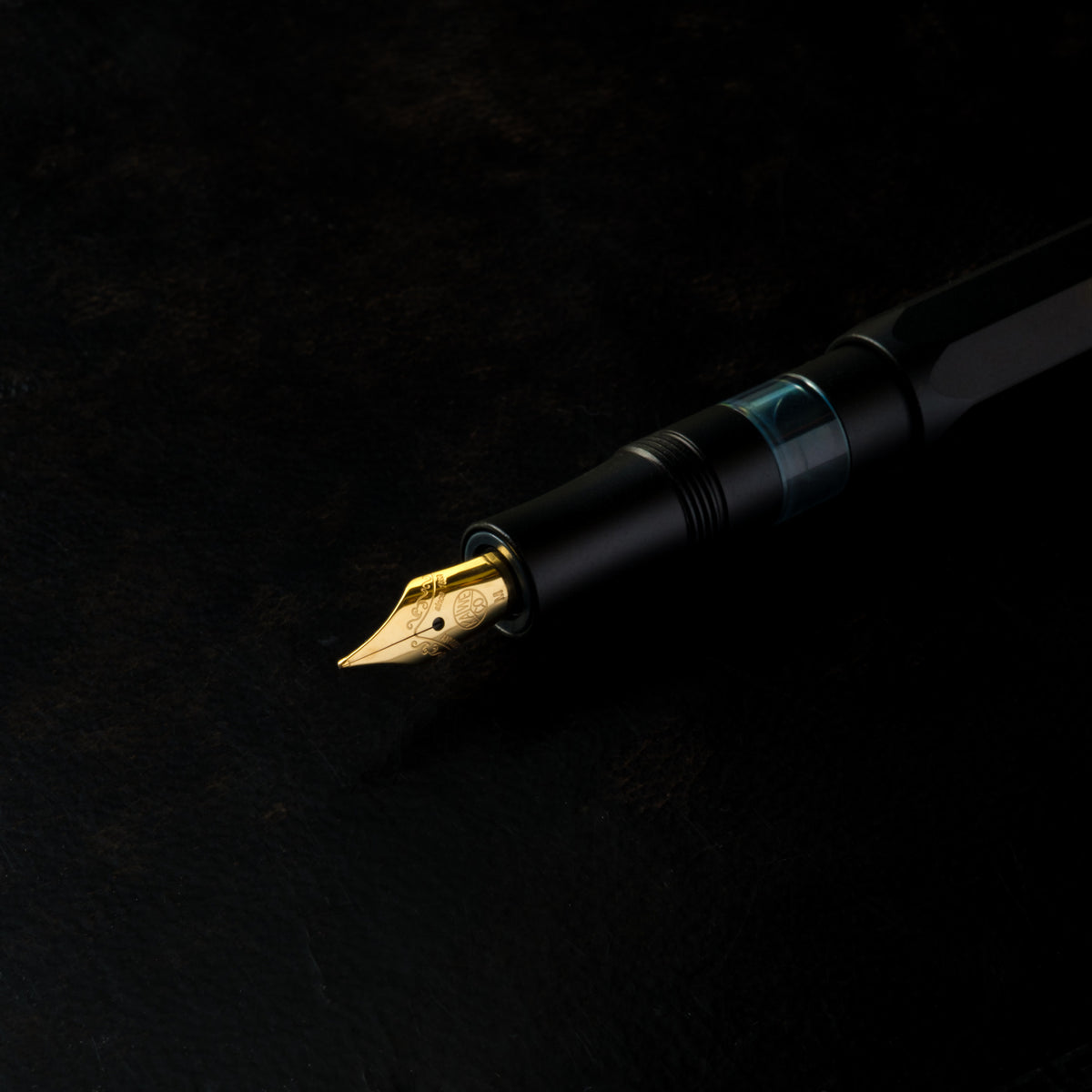 A close-up of Kaweco Piston Sport AL black fountain pen with a gold nib and transparent ink window, resting diagonally on a dark textured background.