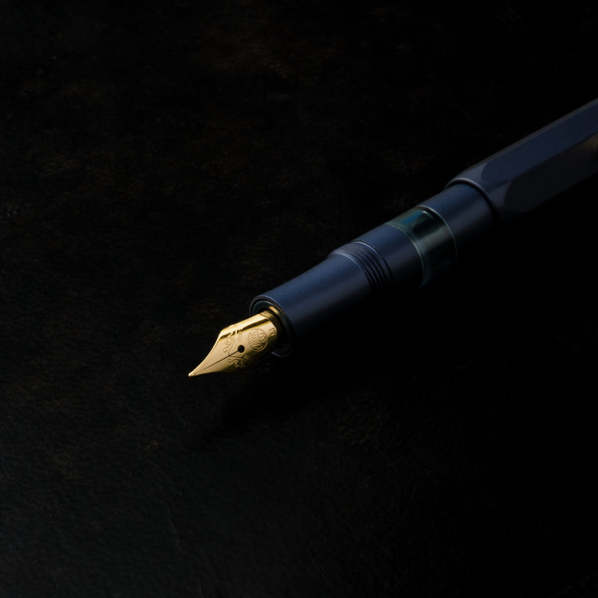 A close-up of Kaweco Piston Sport AL   metallic fountain pen gold nib, resting diagonally on a textured black background.