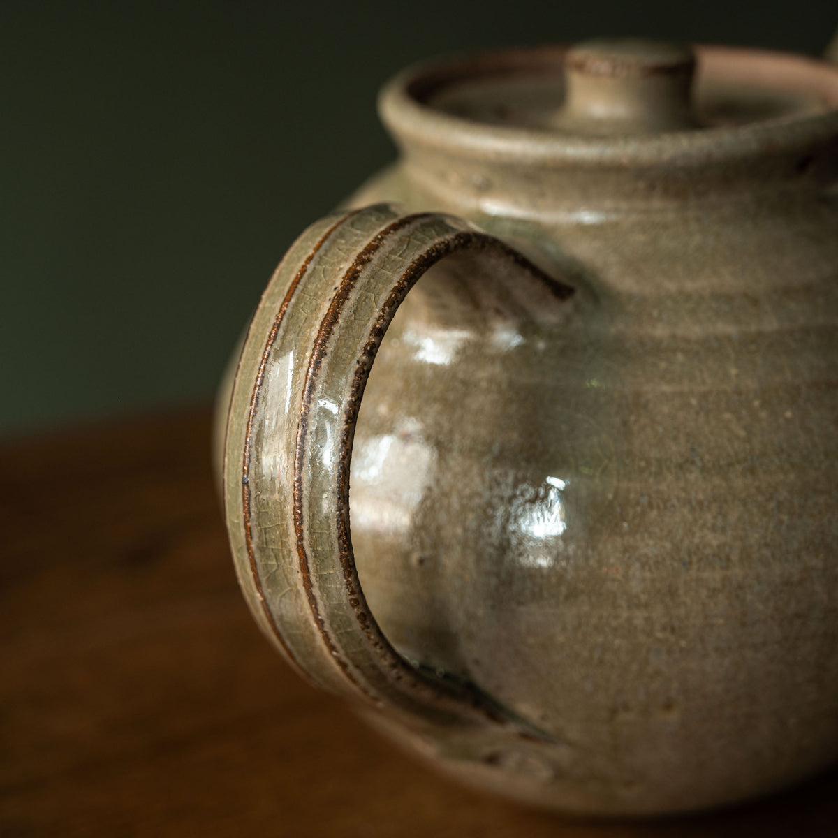Close-up of a handmade Leach Pottery ceramic teapot with a rounded body, curved handle, and matching lid, featuring a glossy earthy Ash green glaze.