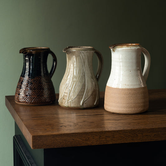 Three handmade Leach Pottery ceramic pitchers with unique glazes and patterns, displayed on a dark wood table against a muted green wall.