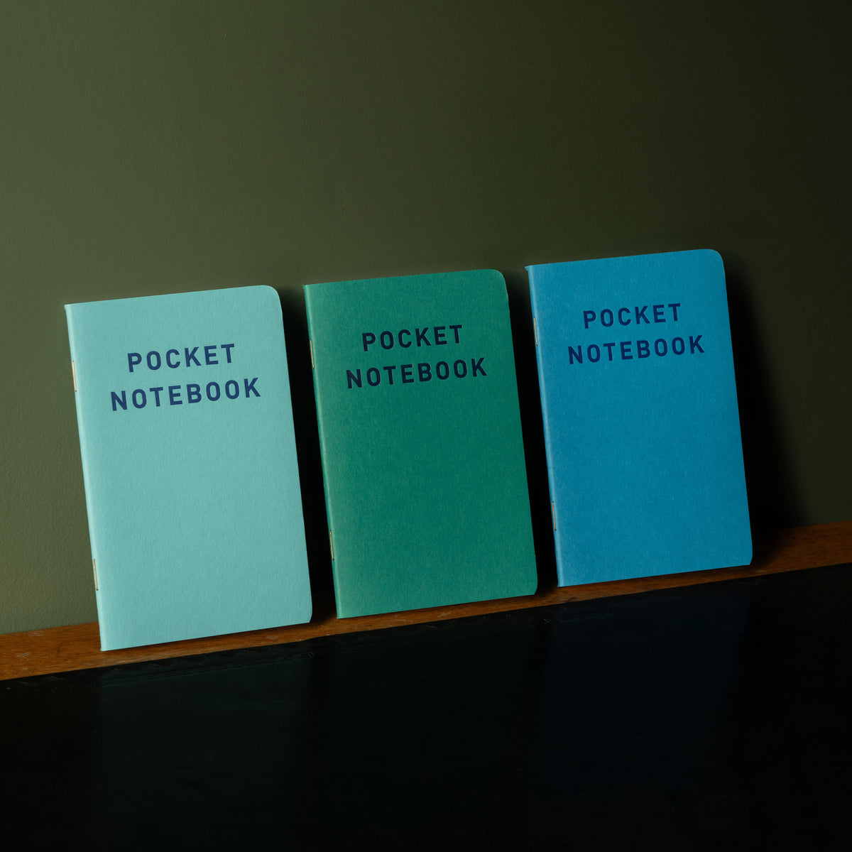 Three pocket notebooks with 'Pocket Notebook' text on a wooden surface against a dark background