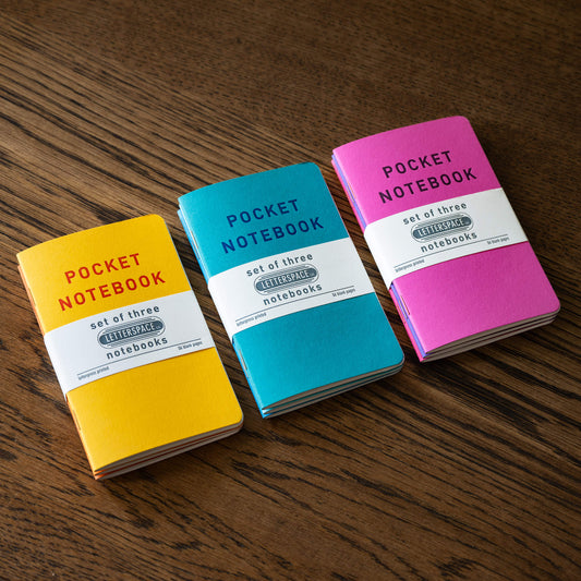 Three small pocket notebook sets in yellow, turquoise and pink are arranged on a dark wood surface. Each notebook has 'POCKET NOTEBOOK' printed on the cover with a white paper band around the middle.