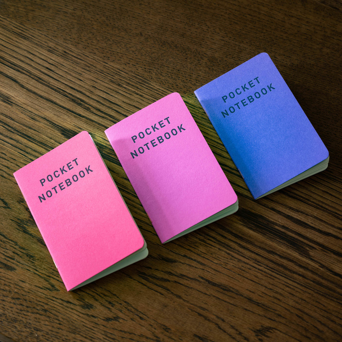 Three small pocket notebooks in pink, purple, and blue are arranged diagonally on a dark wood tabletop. Each notebook has 'POCKET NOTEBOOK' printed on the cover.