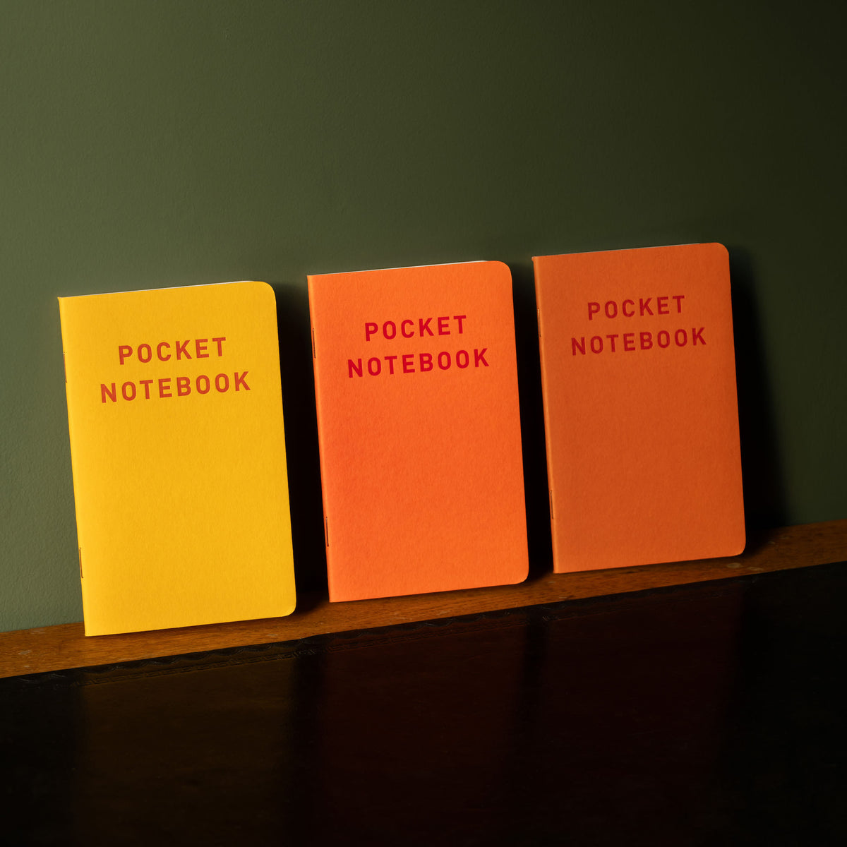Three pocket notebooks in yellow, orange, and red on a wooden shelf against a dark green background.