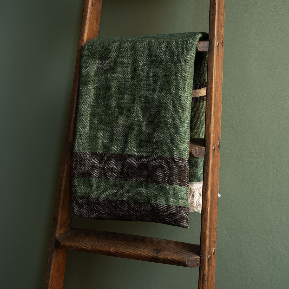 Libeco Belgian Linen Towel. A racing green washed throw with white fringe, draped over a rustic wooden ladder against a dark green wall.