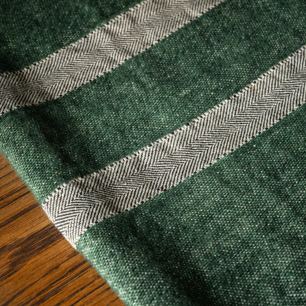 A close-up of Libeco Belgian Linen Towel. With a dark racing green base and two herringbone-striped bands in bone and grey, laid over a wooden surface.