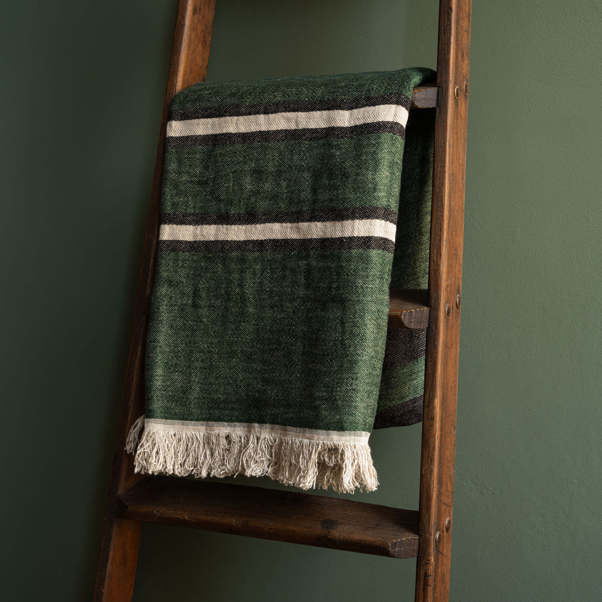 Libeco Belgian Linen Towel. A racing green woven linen throw with bone and black horizontal stripes and fringed tassels, draped over a rustic wooden ladder against a dark green wall.