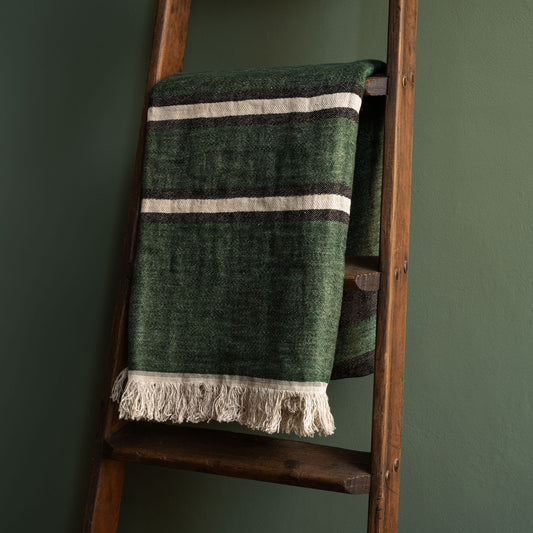 Libeco Belgian Linen Towel. A racing green woven linen throw with bone and black horizontal stripes and fringed tassels, draped over a rustic wooden ladder against a dark green wall.