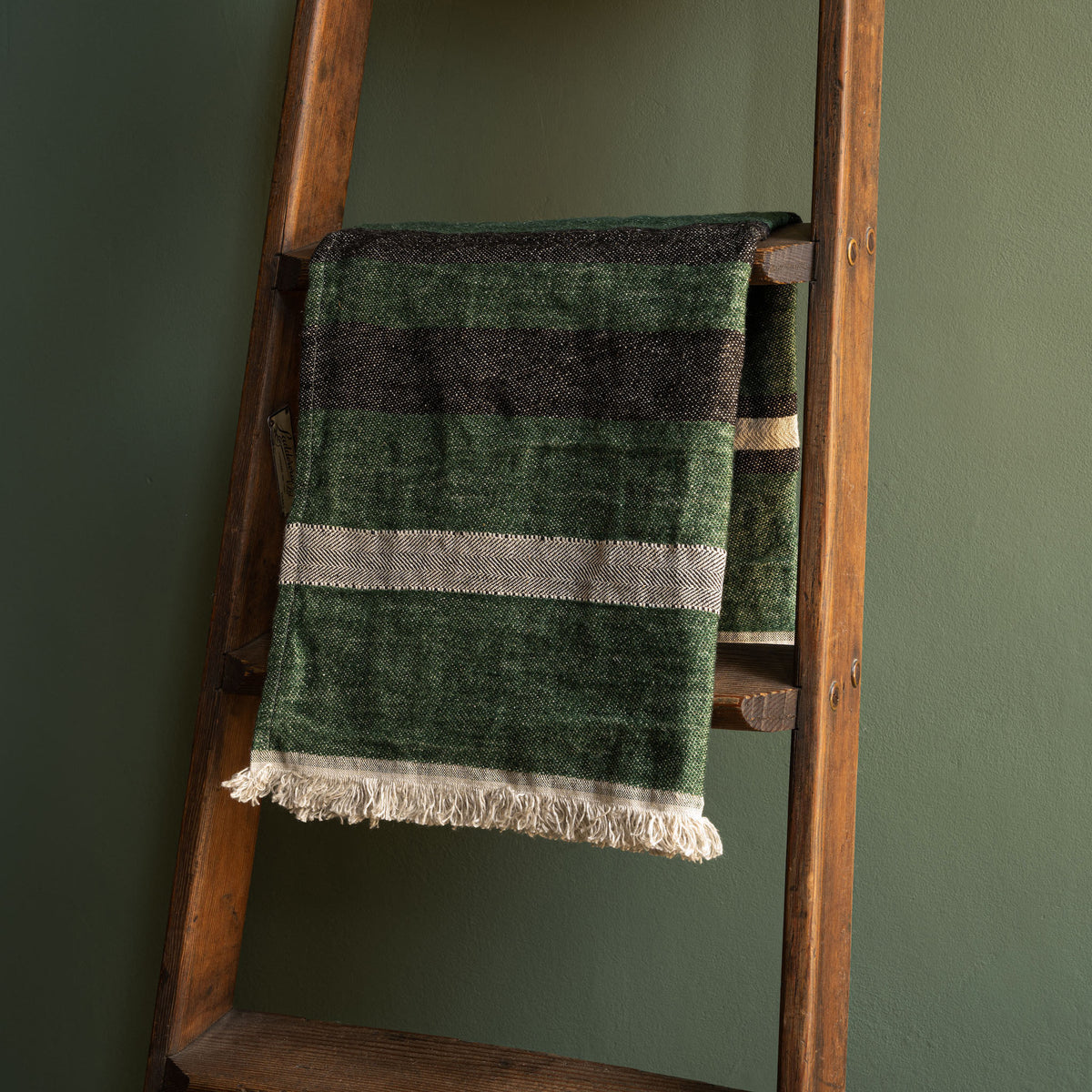 Libeco Belgian Linen Guest Towel. A washed linen hand towel in racing green with black and bone stripes, draped over a rustic wooden ladder against a dark green wall.