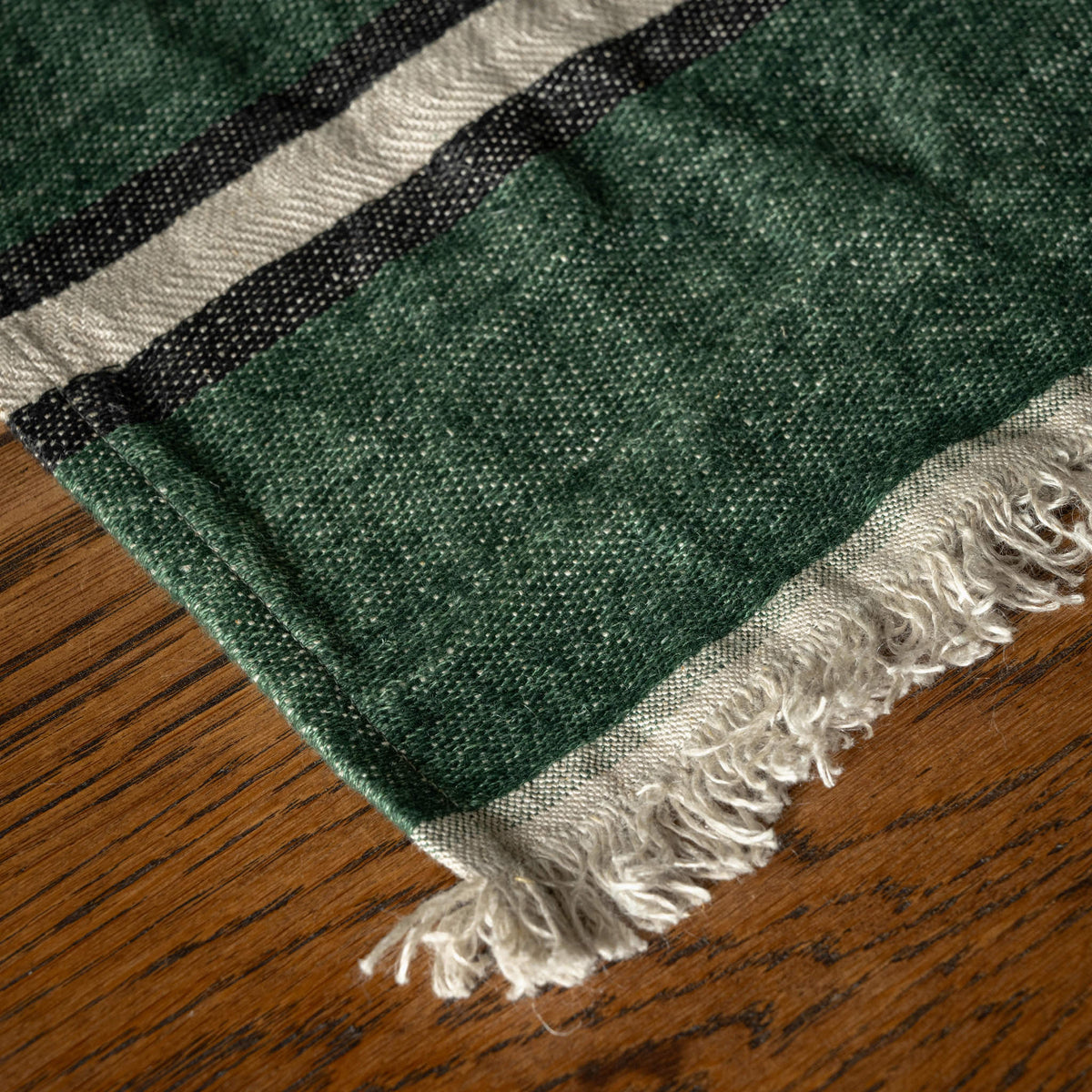 A close-up of Libeco Belgian Linen racing green guest towel. Featuring a racing green body with a cream and black striped border and fringed edges, laid flat on a wooden surface.