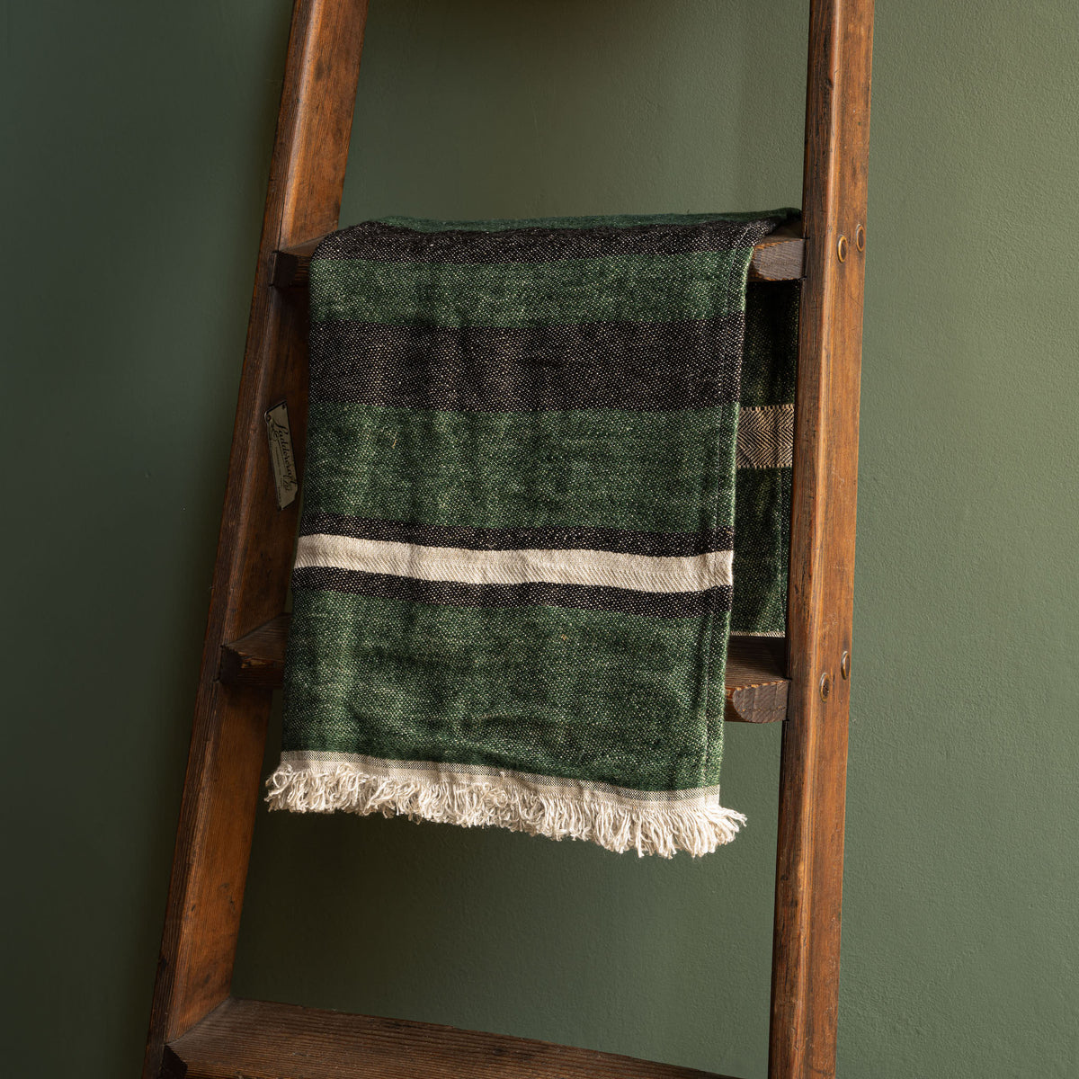 Libeco Belgian Linen Guest Towel. A washed linen racing green hand towel with horizontal stripes in black and bone, draped over a rustic wooden ladder against a dark green wall.