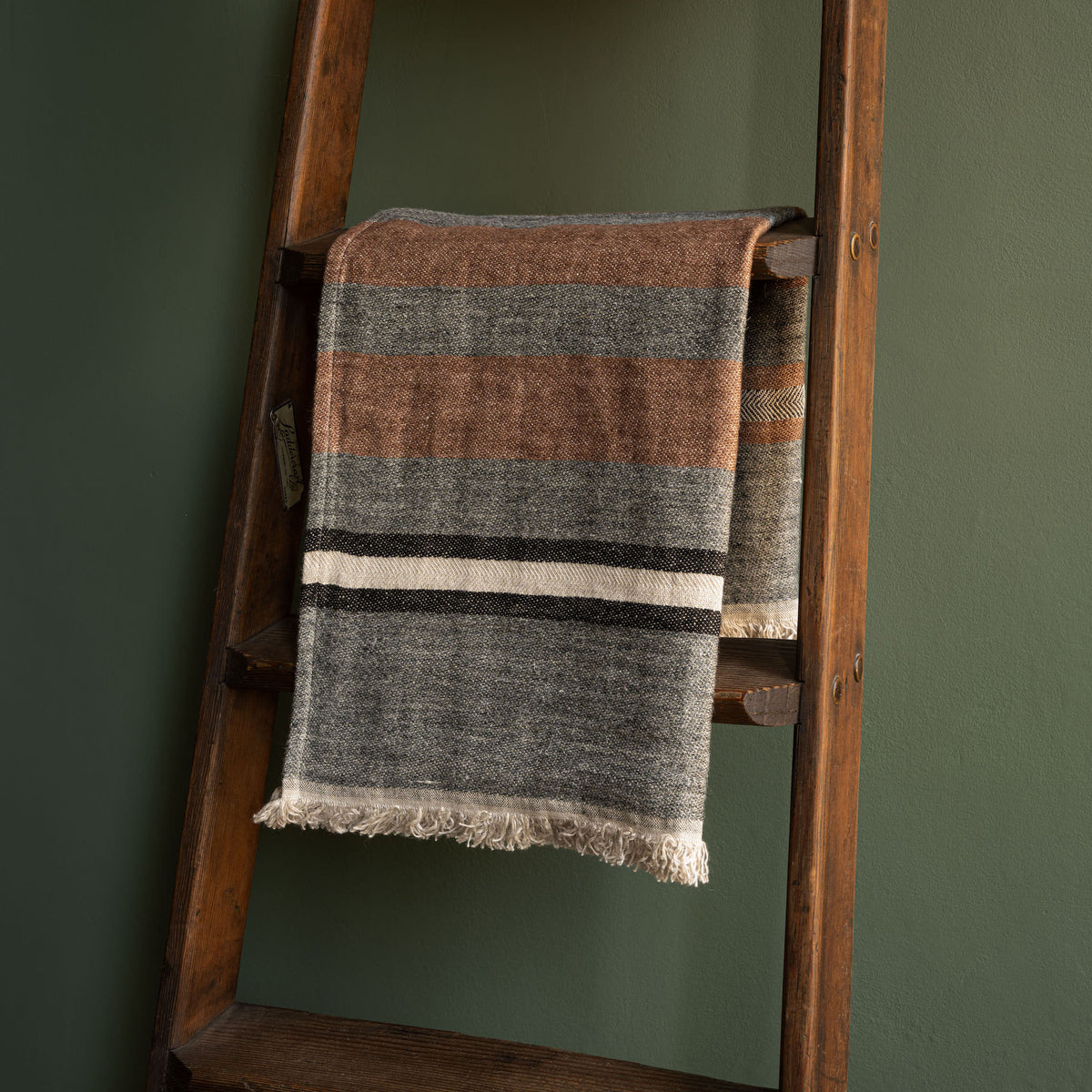 Libeco Belgian Linen Guest Towel. A striped washed linen hand towel with fringed edges draped over a rustic wooden ladder against a dark green wall.