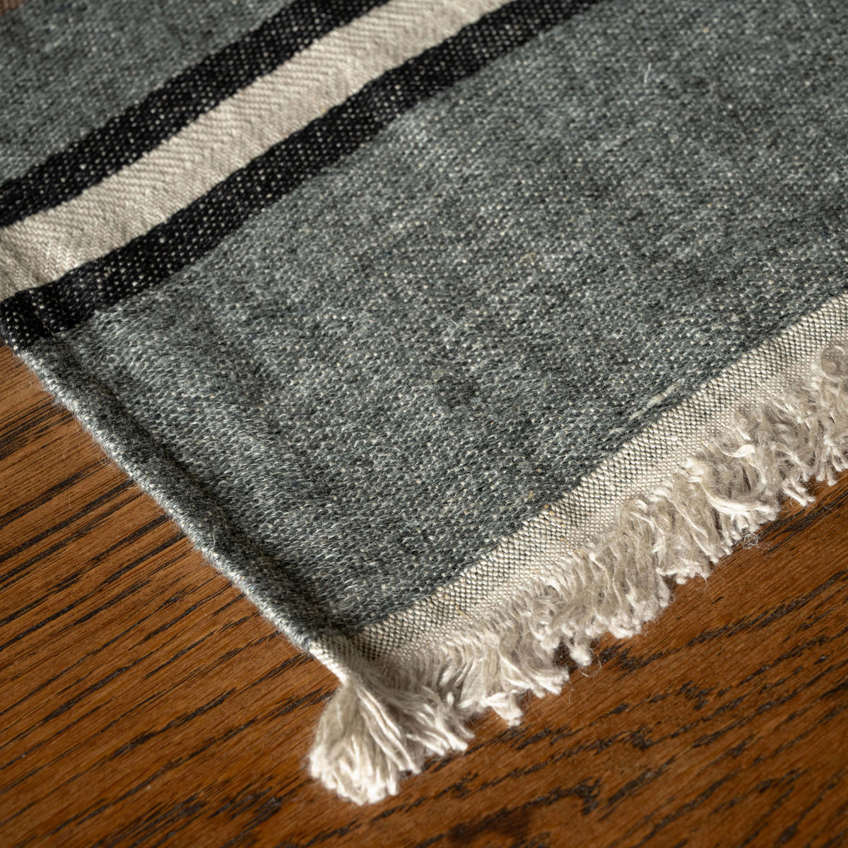 A close-up of Libeco Belgian Linen Guest Towel with fringe, showing a heather grey body with two horizontal cream stripes and a thin black stripe, laid on a brown wooden surface.