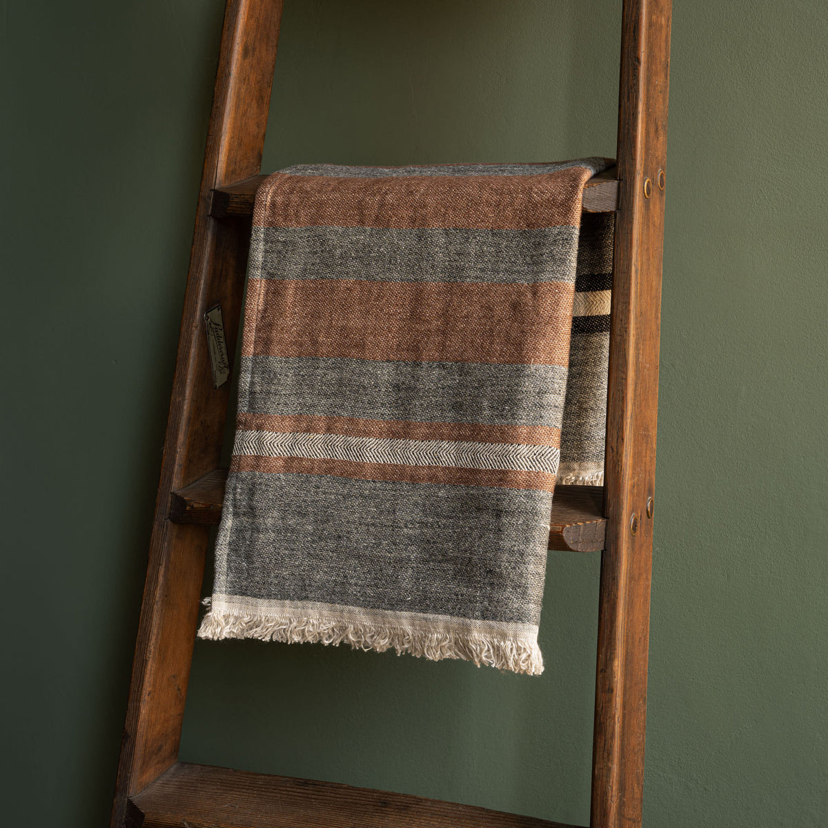 Libeco Belgian Linen Guest Towel. A striped woven hand towel in heather grey with sienna-brown and cream tones, draped over a rustic wooden ladder against a dark green wall.