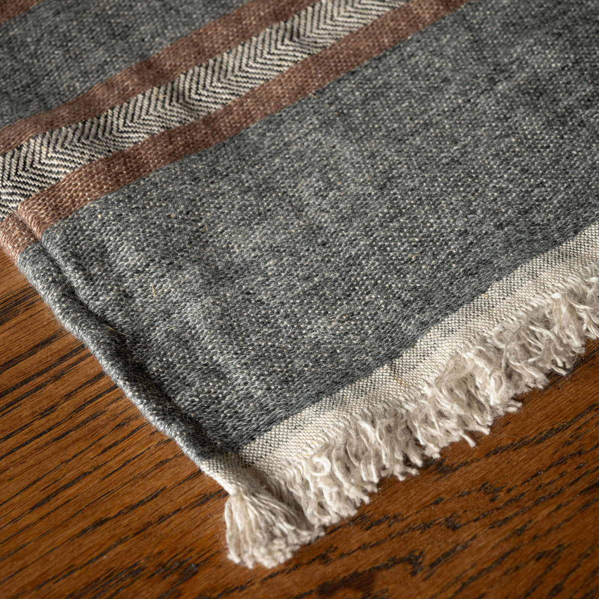 A close-up Libeco Belgian Linen Guest Towel with a heather grey base, rust brown stripes, and off-white fringe, laid on a wooden surface.