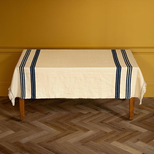 Libeco Belgian Linen 'Indigo Stripe Pièce di Lin'. An off-white woven tablecloth with fringe and dark navy blue stripes, draped over a wooden table against a mustard yellow wall and herringbone wood floor.