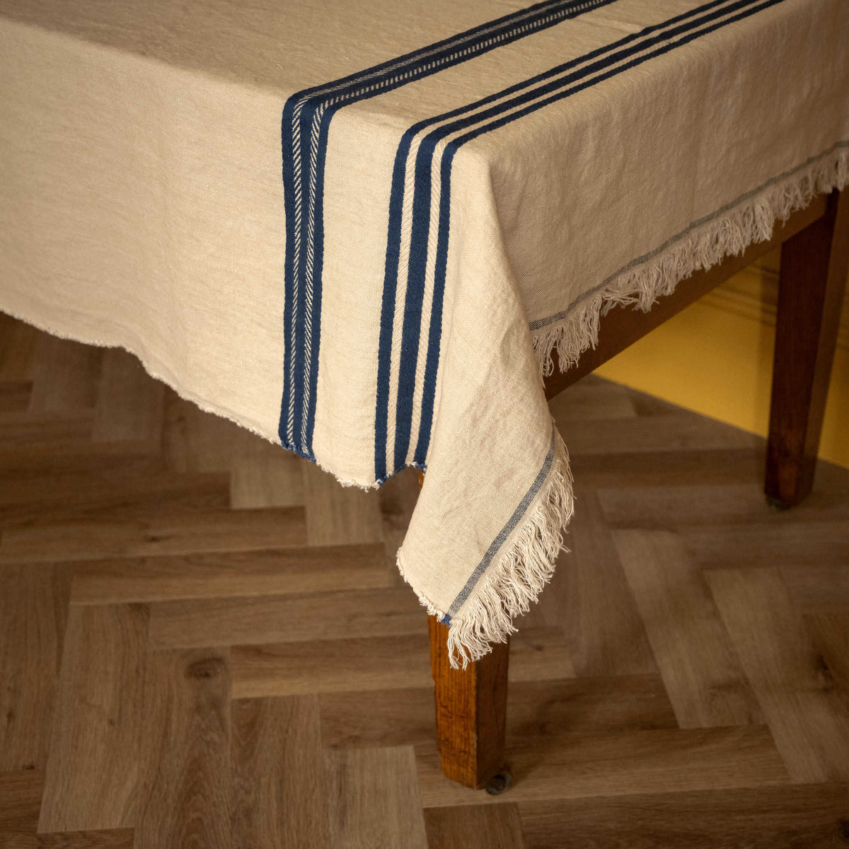Close up of Libeco Belgian Linen 'Pièce di Lin'. An off-white woven tablecloth with dark blue stripes and fringed edges draped over a wooden table, set on a light wood parquet floor.