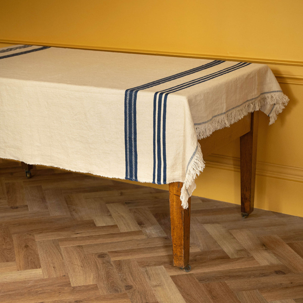 Libeco Belgian Linen Indigo Stripe 'Pièce di Lin'. An off-white coloured tablecloth with blue stripes and fringe trim draped over a wooden table in a room with a mustard yellow wall and herringbone wood floor.