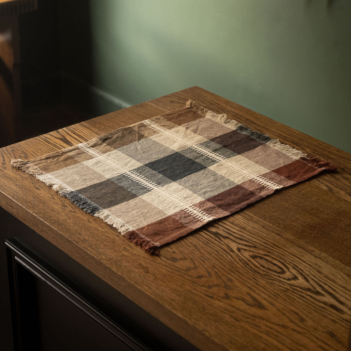Libeco Belgian Linen Napkin. A square plaid fringed linen napkin or placemat in brown, beige, and grey tones, laid on a dark wood table.