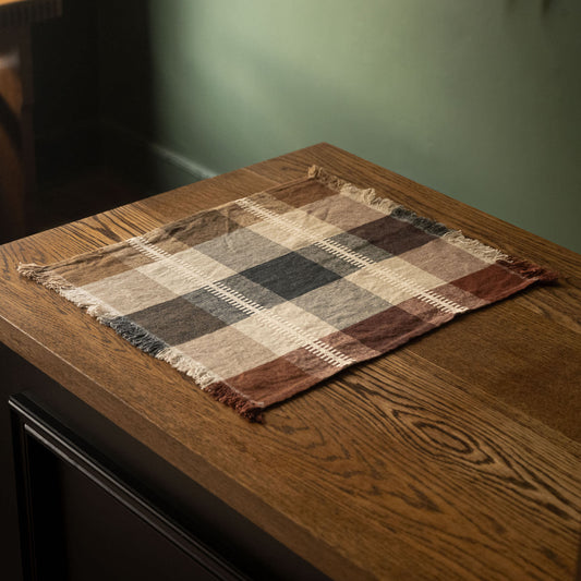 Libeco Belgian Linen Napkin. A square plaid fringed linen napkin or placemat in brown, beige, and grey tones, laid on a dark wood table.