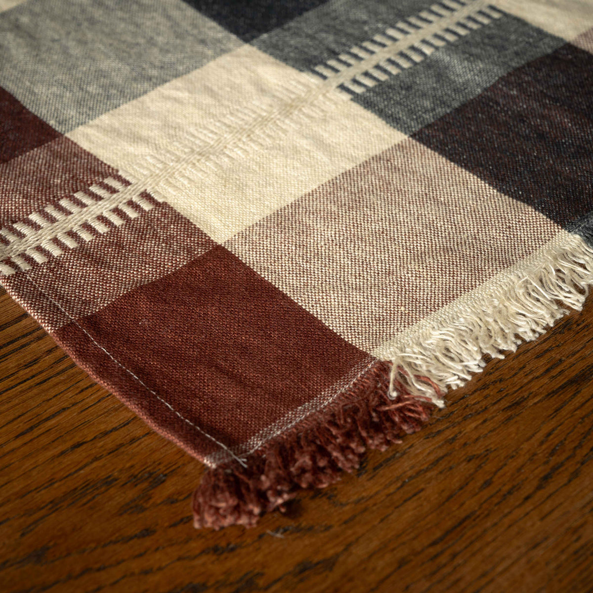 A close-up of Libeco Linen Maxine Napkin. A plaid woven napkin with fringed edges, laid on a wooden surface. The pattern includes cream, brown and taupe blocks, with visible stitched stripe details along the border.