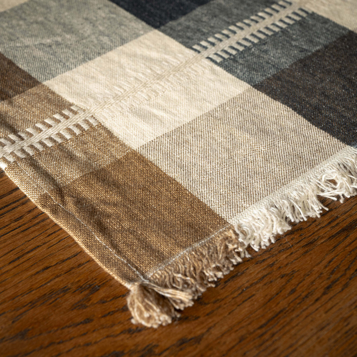 A close-up of Libeco Belgian Linen Maxine Napkin. A washed woven linen napkin features a check pattern in beige, brown, cream, and dark grey, with a textured horizontal stripe detail.with fringe, laid on a dark wood surface. 
