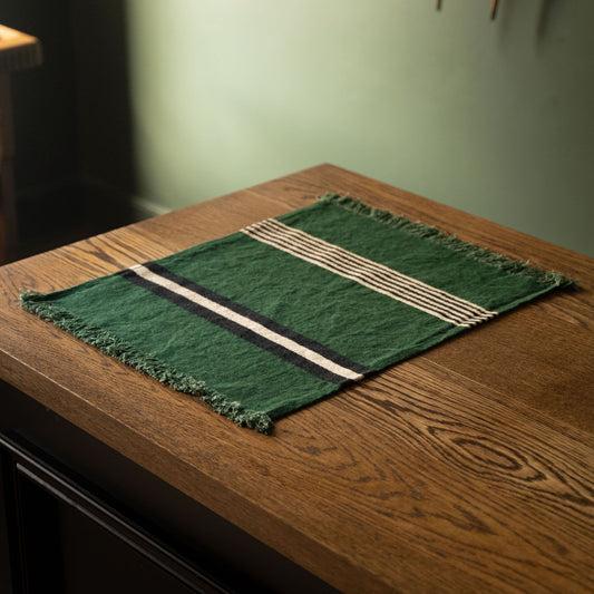 Libeco Belgian Linen 'Nell' Napkin / Placemat. A racing green woven linen placemat or napkin with fringe edges and striped detailing, displayed on a wooden table in a dimly lit interior setting.