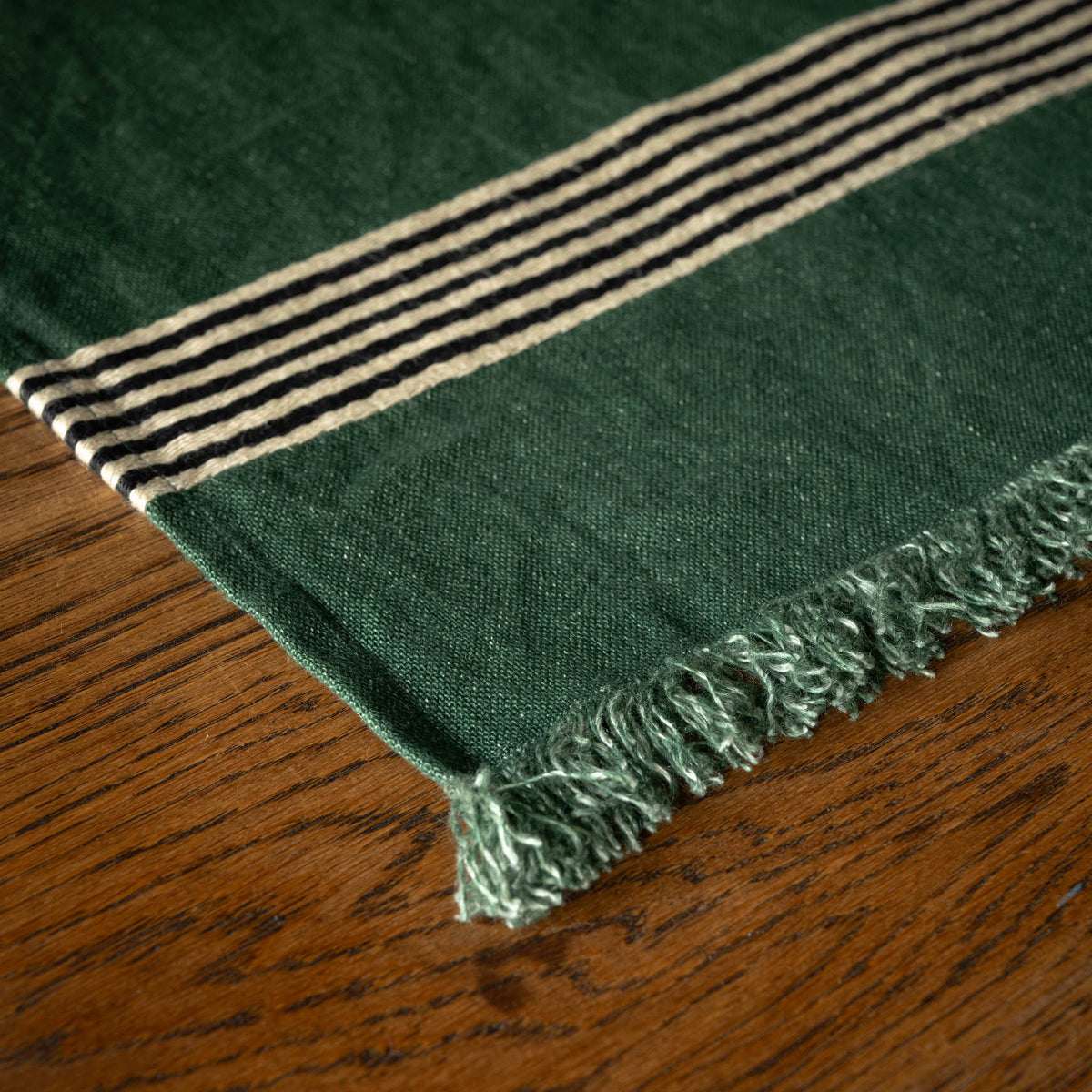 A close-up of Libeco Belgian Linen 'Nell' Napkin / Placemat. A dark green woven textile with fringe and a striped border, laid on a brown wooden surface.