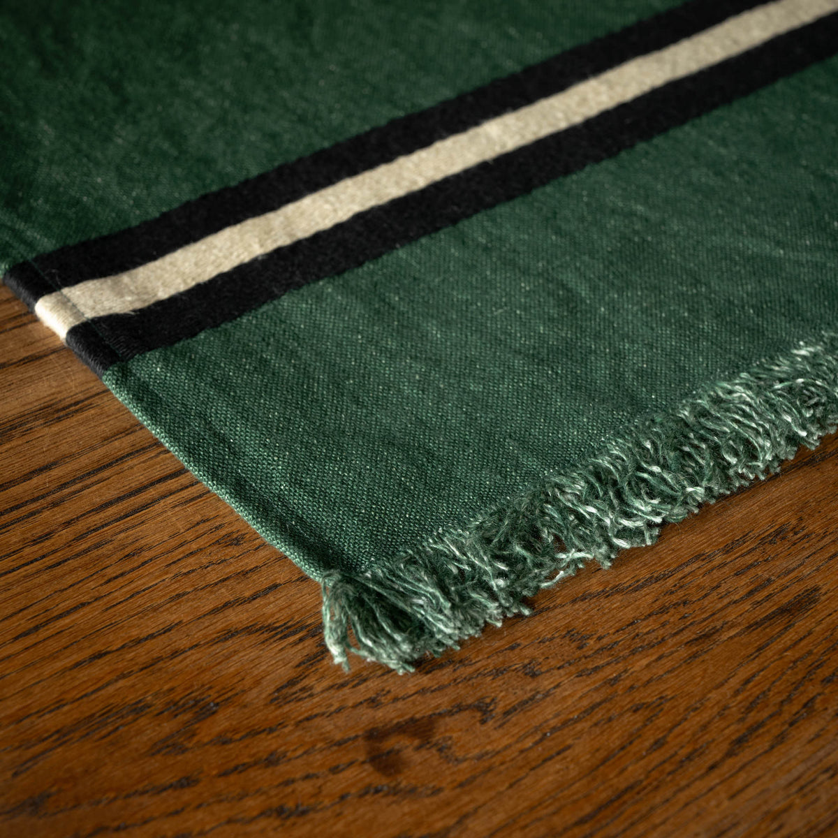 A close-up Libeco Belgian Linen 'Nell' napkin / placement. Dark racing green woven base with fringe edge, featuring two horizontal stripes in black and cream, laid on a wooden table.