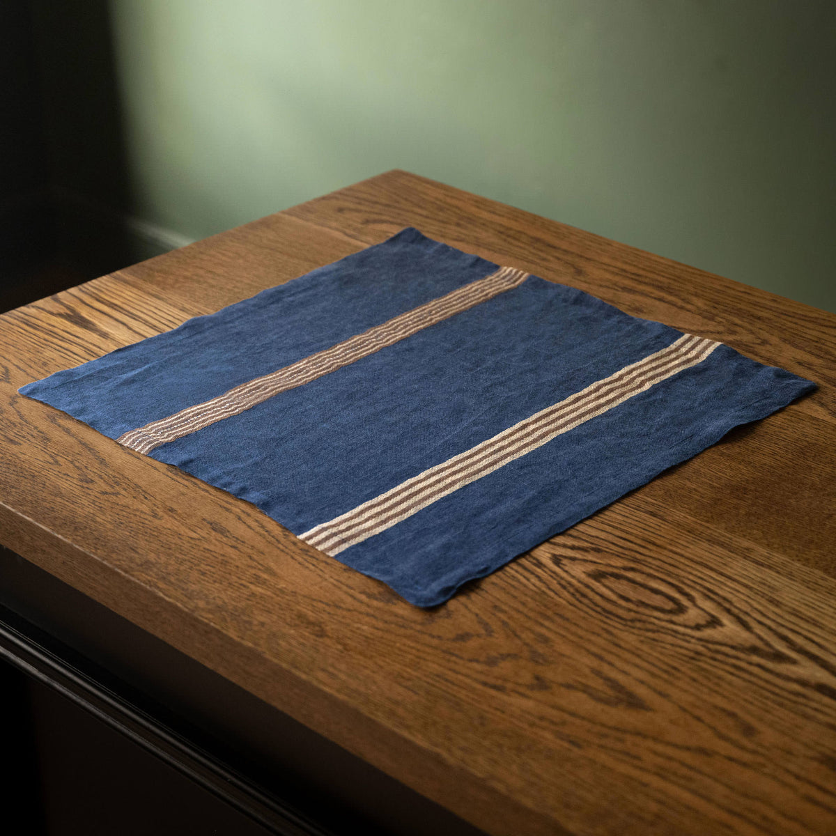 Libeco Belgian Linen 'Noa' Napkin. A square indigo blue open weave  napkin with two beige & brown stripes, laid out on a rustic wooden table.