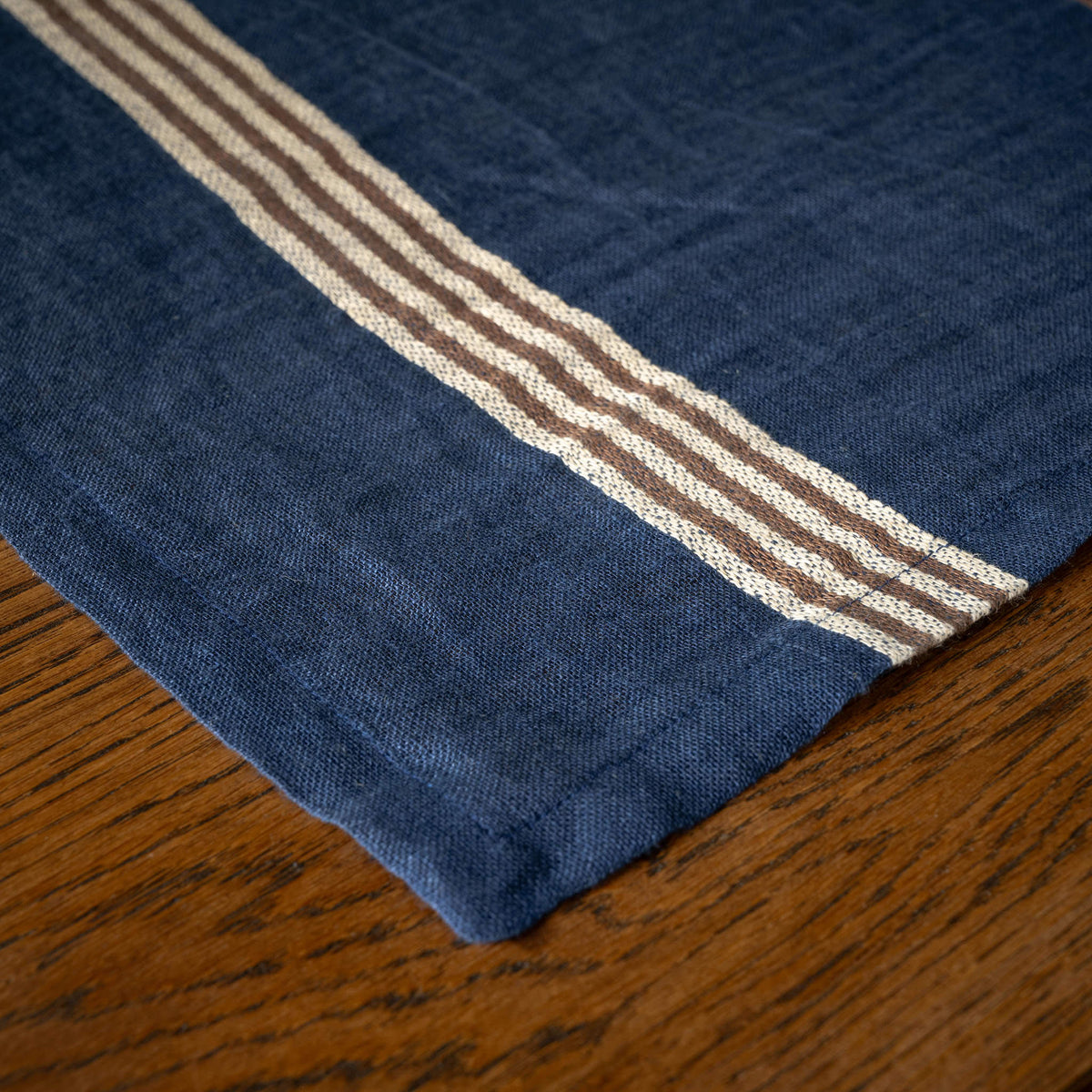 A close-up of Libeco Belgian Linen 'Noa' Napkin. Indigo blue base, featuring two horizontal  stripes in ecru and brown, laid on a wooden table.