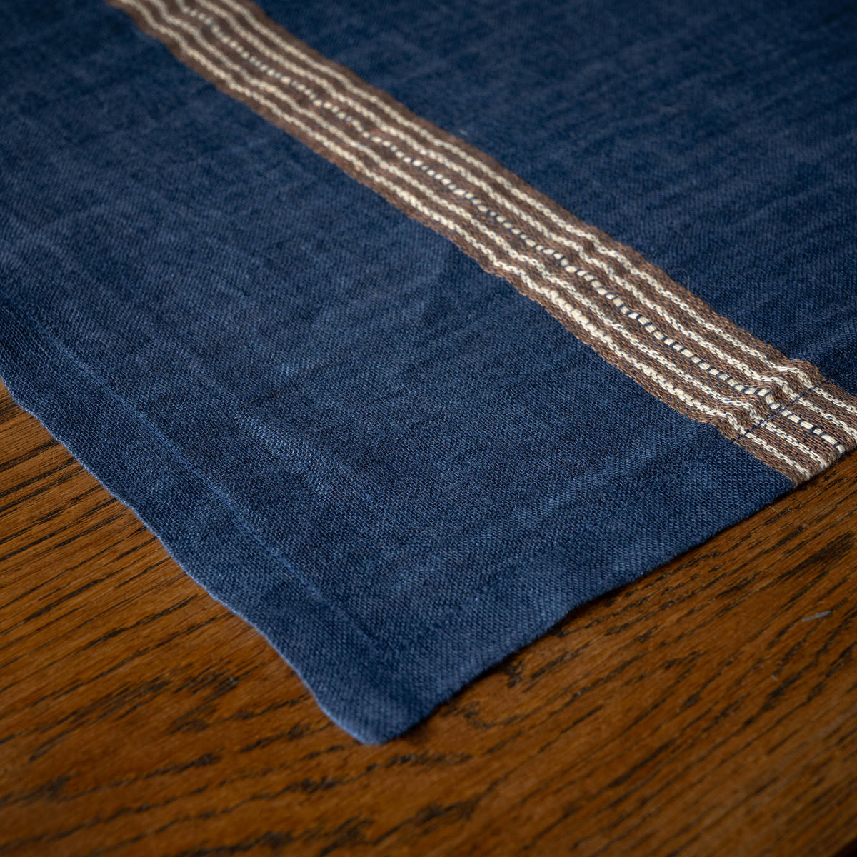 A close-up of Libeco Belgian Linen 'Noa' Napkin. Indigo blue base featuring ecru and brown stripes, laid on a wooden table.