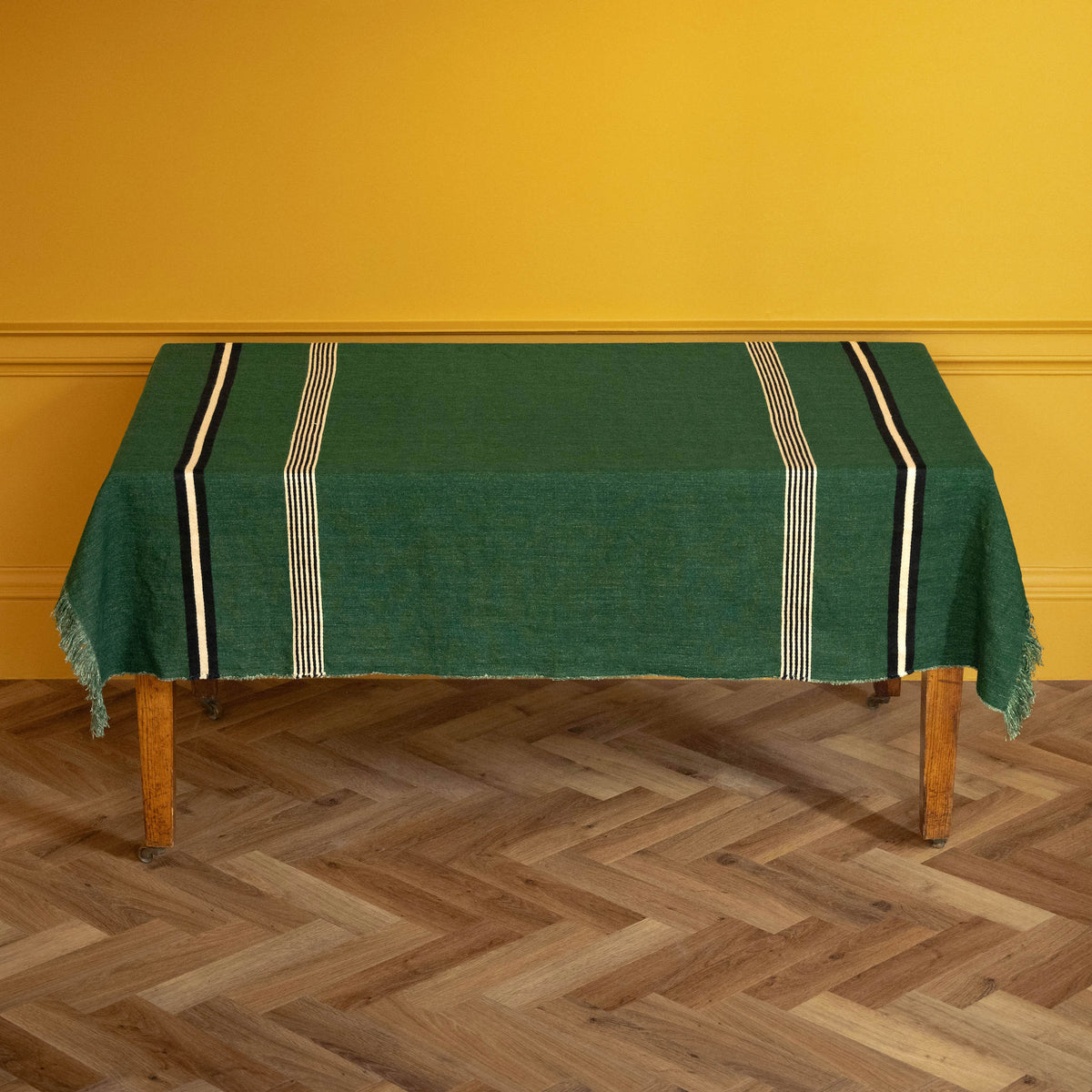 Libeco Belgian Linen 'Nell" Pièce di Lin. A racing green woven linen tablecloth with cream and black striped borders and fringe, draped over a wooden table against a mustard yellow wall and herringbone wood floor.