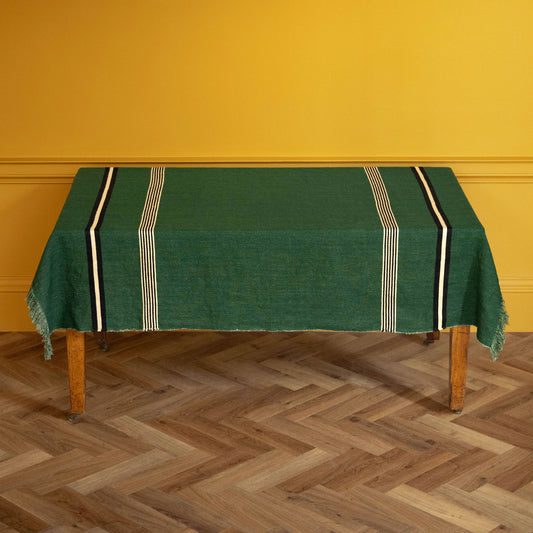 Libeco Belgian Linen 'Nell" Pièce di Lin. A racing green woven linen tablecloth with cream and black striped borders and fringe, draped over a wooden table against a mustard yellow wall and herringbone wood floor.
