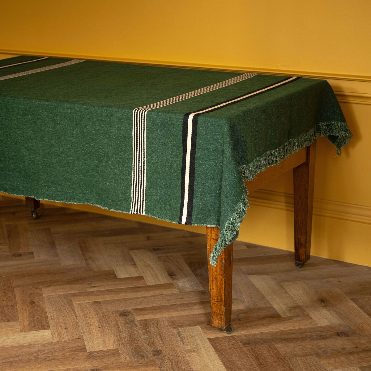 Libeco Belgian Linen 'Nell" Pièce di Lin. A dark green woven linen tablecloth with fringe and striped accents draped over a wooden table in a room with a mustard yellow wall and herringbone wood floor.